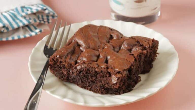 Um pedaço de brownie com textura macia e crocante na superfície, servido em um prato branco com um garfo ao lado