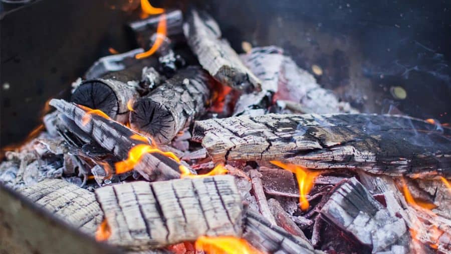 Troncos de madeira parcialmente queimados em uma fogueira, com chamas alaranjadas e cinzas ao redor