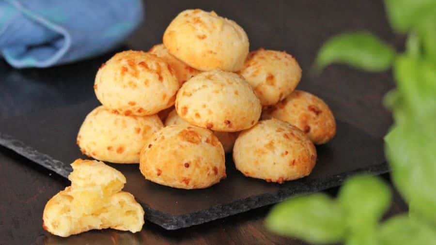 Pão de queijo com casca dourada e textura rústica, empilhado sobre uma base preta, com um pedaço mordido ao lado