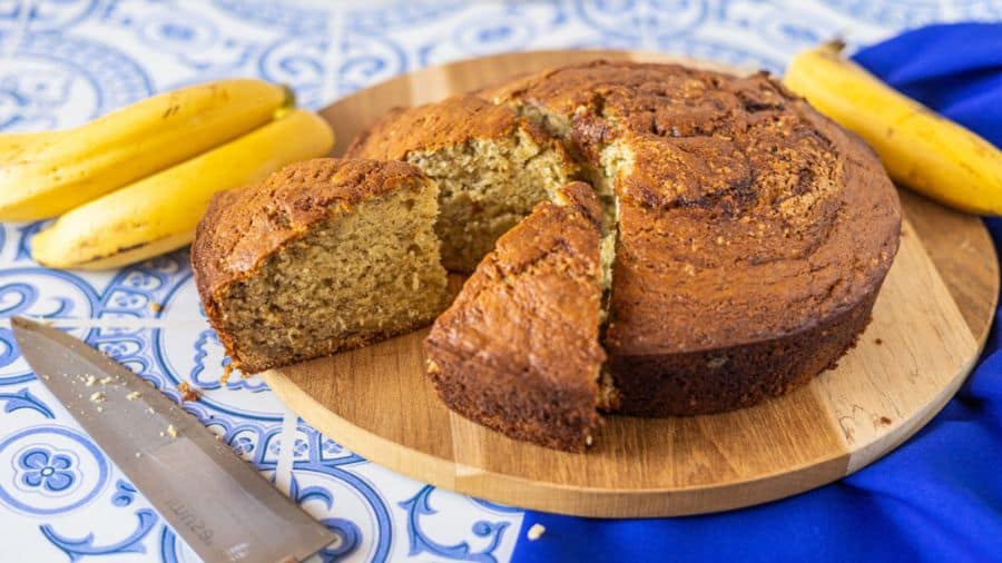 Bolo de banana redondo com fatias cortadas sobre uma tábua de madeira, decorado com bananas ao fundo em uma bancada com azulejos azuis