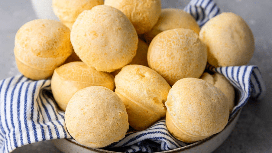 Pão de queijo tradicional com superfície lisa e dourada, servido em uma tigela forrada com pano listrado azul e branco