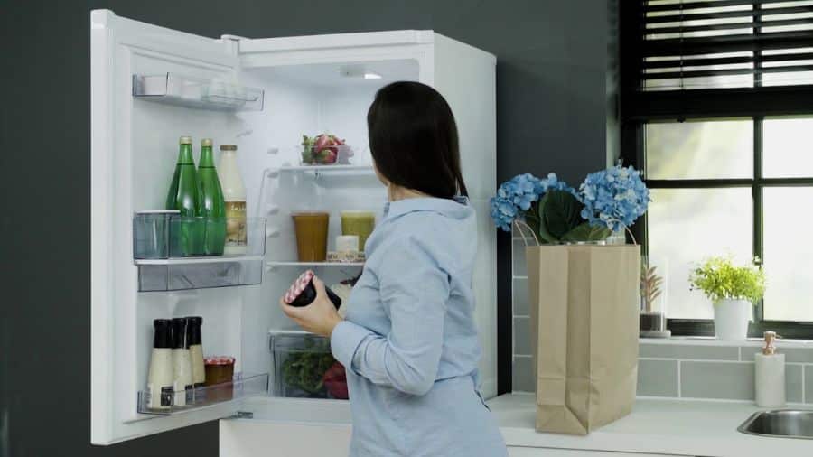 Mulher ajustando a temperatura interna da geladeira com a porta aberta, segurando um pote e observando o interior do refrigerador