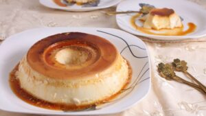 Pudim de leite condensado com calda caramelizada servido em prato branco, com uma fatia separada ao fundo sobre a mesa