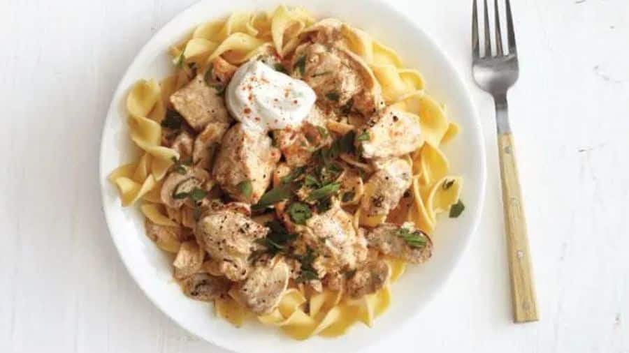 Strogonoff de frango com molho cremoso, servido sobre macarrão tipo fita e decorado com creme azedo e salsa