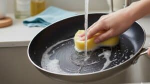 Pessoa lavando frigideira antiaderente com esponja sob água corrente, usada como imagem destacada do artigo