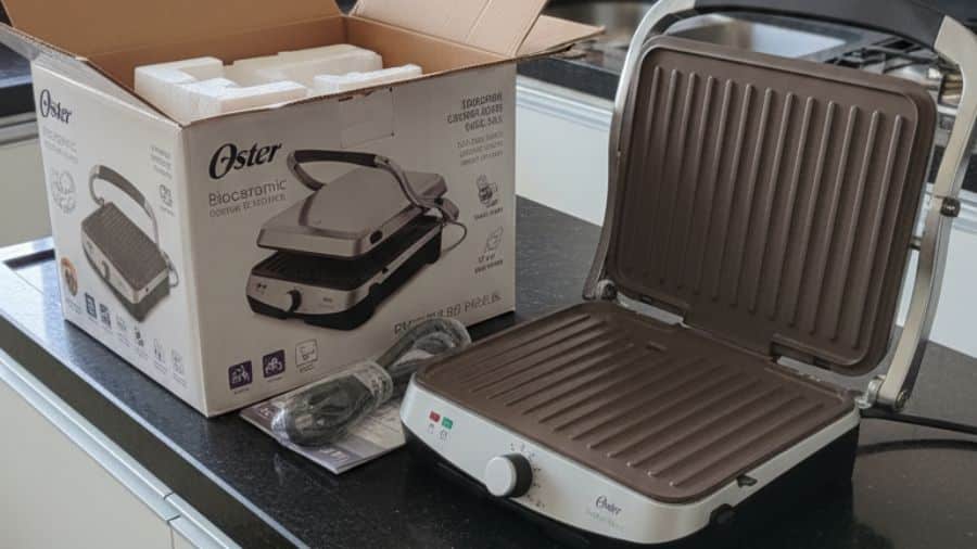 Grill Oster Bioceramic 2 em 1 aberto ao lado da caixa e acessórios sobre bancada de cozinha moderna.