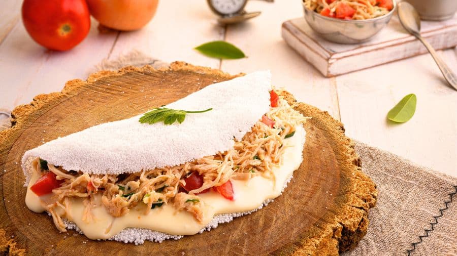 Tapioca aberta com recheio de frango desfiado, tomate picado e queijo derretido sobre base rústica de madeira, usada como imagem ilustrativa do artigo