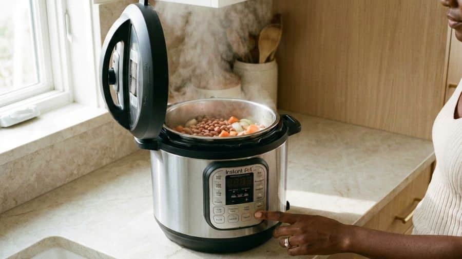 Panela de pressão elétrica com tampa aberta, feijão cozinhando com legumes e botão sendo pressionado por mulher